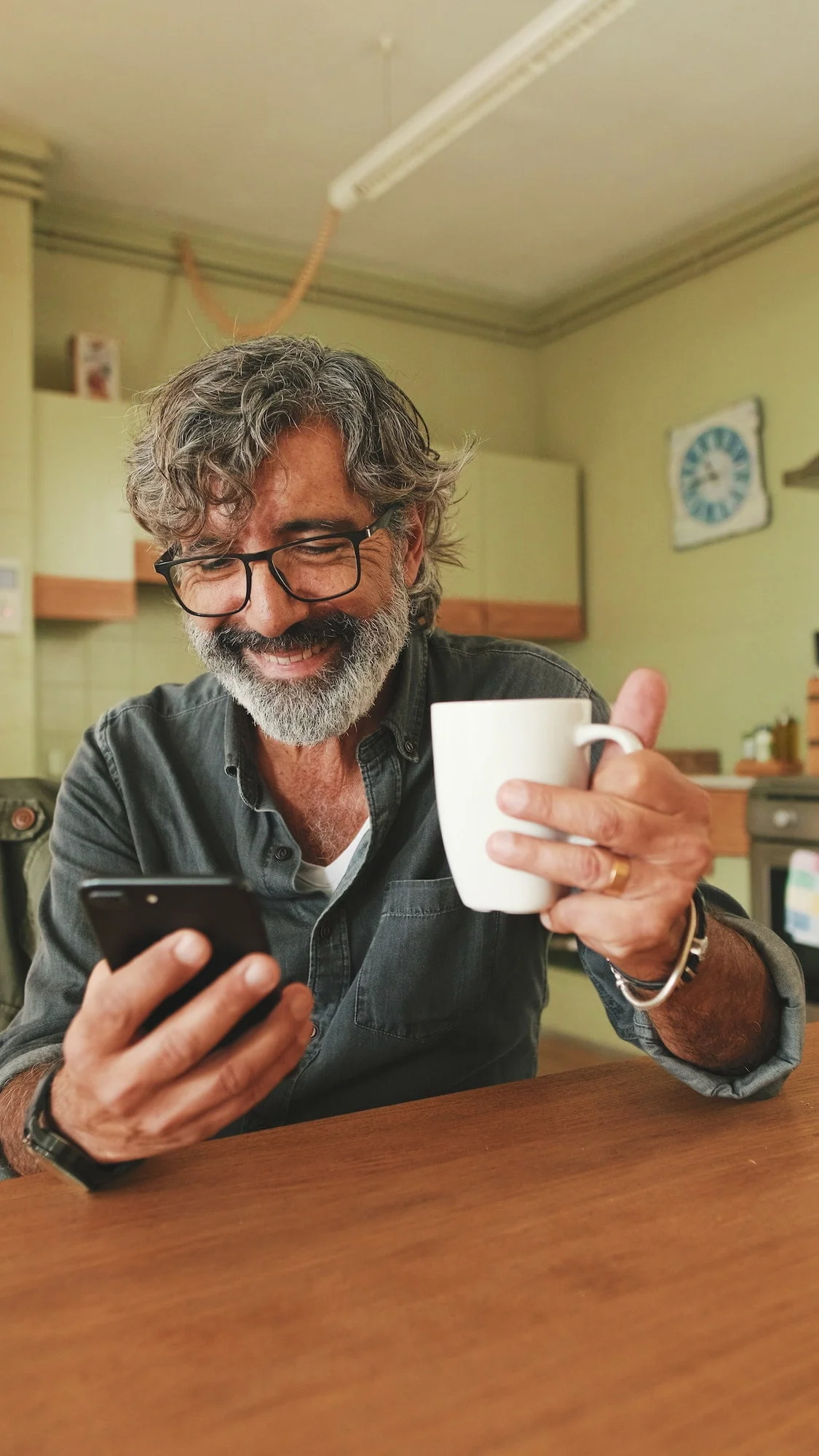 Elderly man drinks coffee from mug and uses mobile phone at home